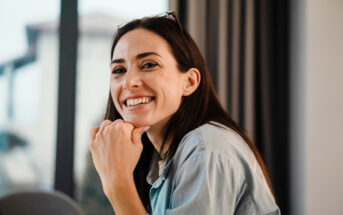 A woman with long dark hair is smiling warmly while resting her chin on her hand. She is wearing a light blue denim shirt and has a pair of glasses perched on her head. The background is softly blurred, showing a window with curtains and a glimpse of an outdoor scene.