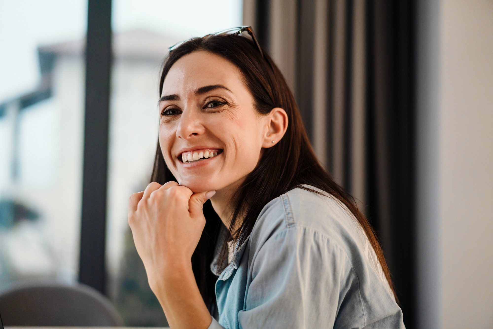 A woman with long dark hair is smiling warmly while resting her chin on her hand. She is wearing a light blue denim shirt and has a pair of glasses perched on her head. The background is softly blurred, showing a window with curtains and a glimpse of an outdoor scene.