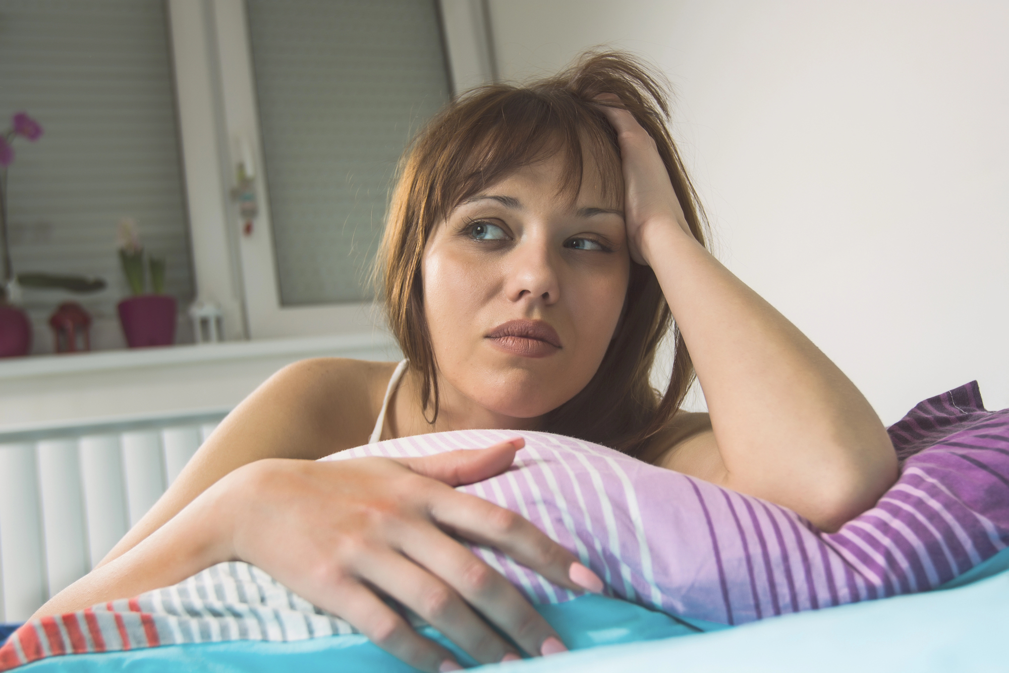 A woman with short brown hair sits on a colorful striped blanket, resting her head on her hand while gazing off to the side. She appears contemplative or possibly concerned. Behind her, there is a window with closed blinds and some potted plants on the windowsill.