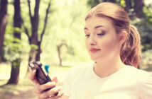 A young woman with a ponytail is outdoors in a park, holding a smartphone in her hand and looking at it with a slightly puzzled expression. She is wearing a white t-shirt, and blurred trees are visible in the background.