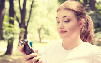 A young woman with a ponytail is outdoors in a park, holding a smartphone in her hand and looking at it with a slightly puzzled expression. She is wearing a white t-shirt, and blurred trees are visible in the background.