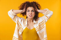 A woman with curly hair pulls at her hair with both hands and displays a frustrated expression. She is wearing a yellow top and a white and orange striped shirt, standing against a bright yellow background.