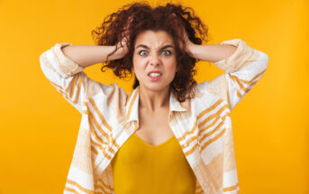 A woman with curly hair pulls at her hair with both hands and displays a frustrated expression. She is wearing a yellow top and a white and orange striped shirt, standing against a bright yellow background.