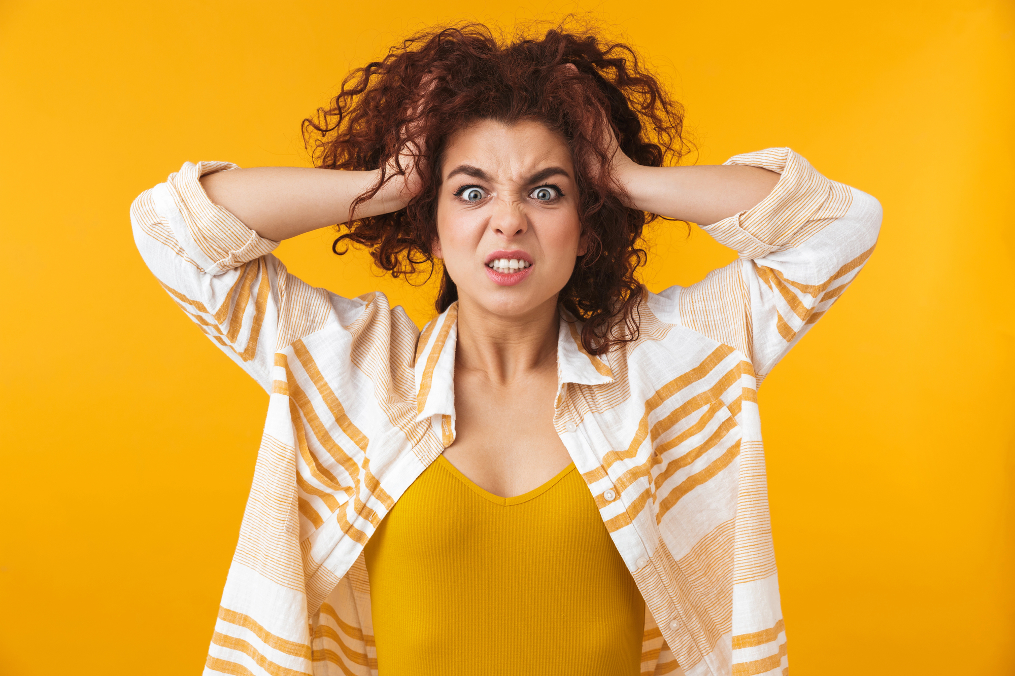 A woman with curly hair pulls at her hair with both hands and displays a frustrated expression. She is wearing a yellow top and a white and orange striped shirt, standing against a bright yellow background.