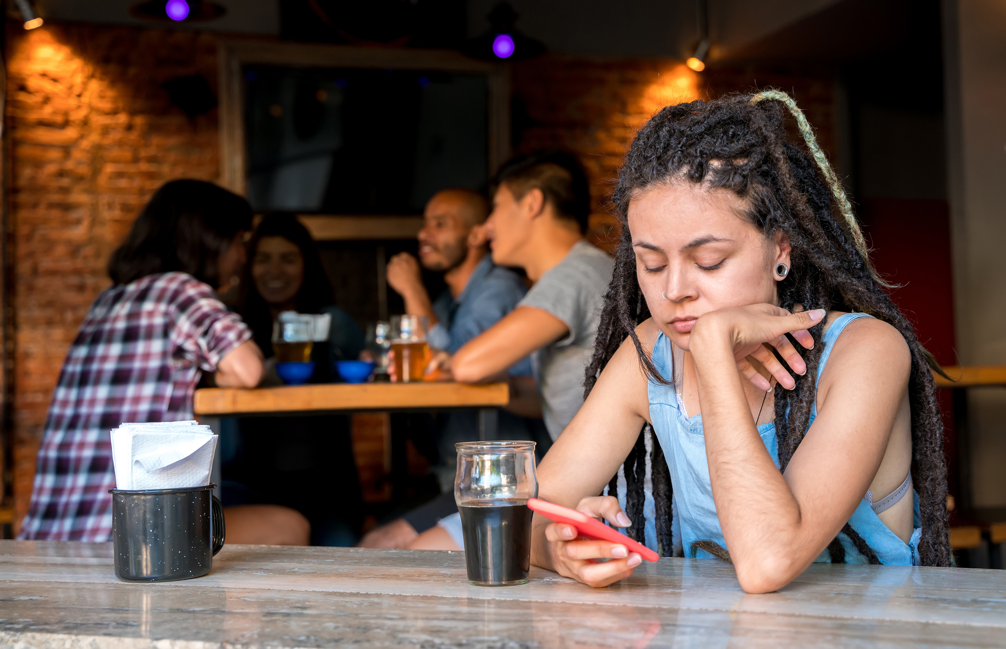 A person with dreadlocks sits alone at a table, looking at their phone. They seem disinterested in the surroundings. In the background, a group of people are engaged in conversation and laughter while drinking at another table.