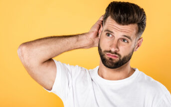 A man with short brown hair and a neatly trimmed beard stands against a solid yellow background. He wears a white t-shirt and has a slightly puzzled expression, scratching the back of his head with one hand.