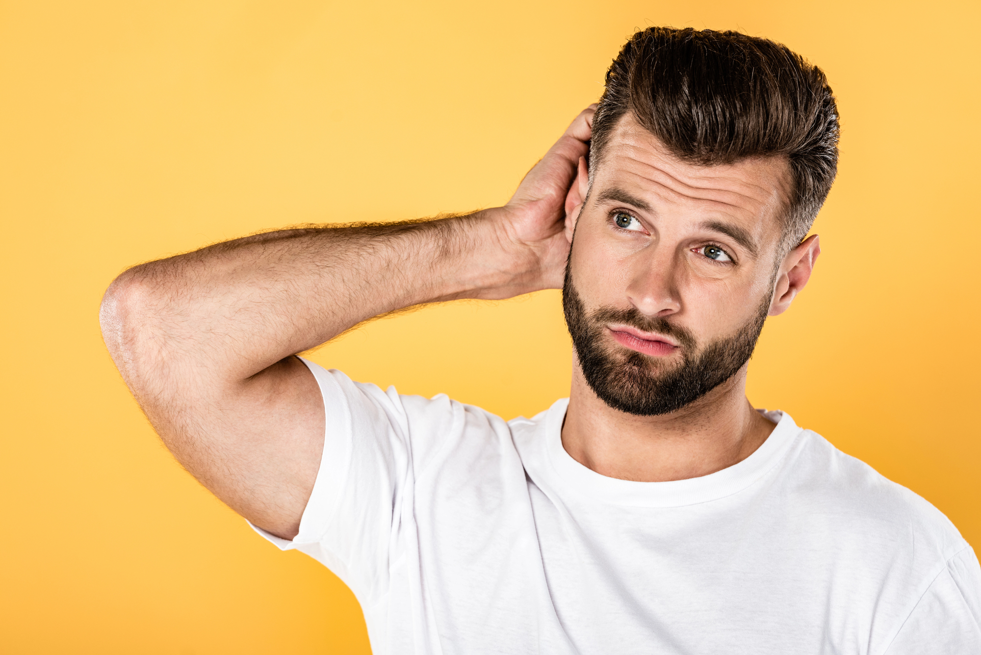 A man with short brown hair and a neatly trimmed beard stands against a solid yellow background. He wears a white t-shirt and has a slightly puzzled expression, scratching the back of his head with one hand.