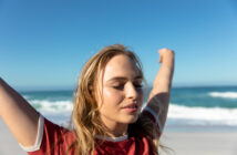 A person with long blonde hair is standing on a beach with eyes closed, arms raised, and a peaceful expression. The person is wearing a red and white T-shirt. The ocean and blue sky serve as the backdrop.