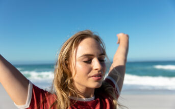 A person with long blonde hair is standing on a beach with eyes closed, arms raised, and a peaceful expression. The person is wearing a red and white T-shirt. The ocean and blue sky serve as the backdrop.