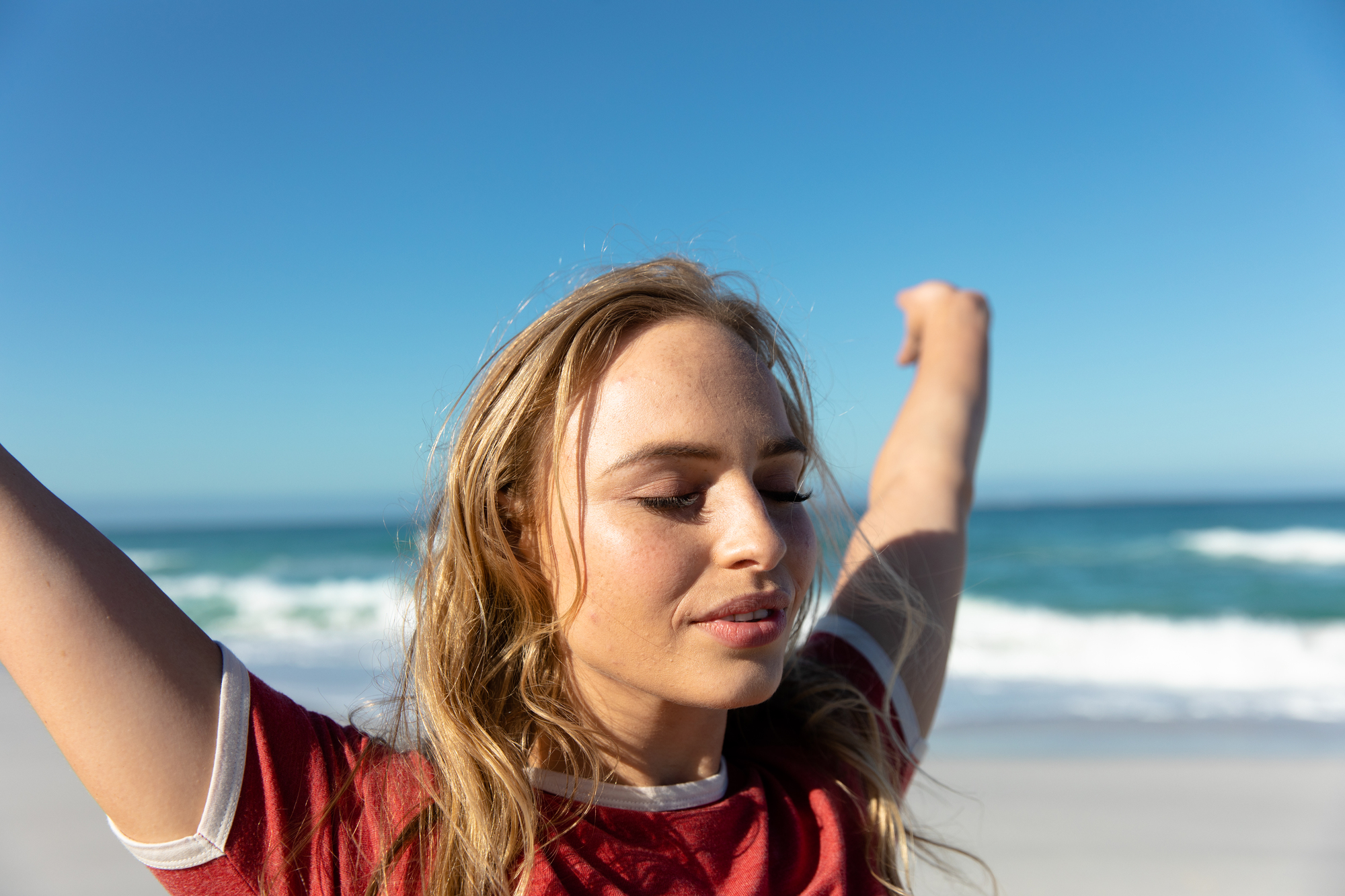 A person with long blonde hair is standing on a beach with eyes closed, arms raised, and a peaceful expression. The person is wearing a red and white T-shirt. The ocean and blue sky serve as the backdrop.