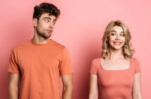 A man with curly dark hair in an orange t-shirt looks sideways at a smiling blonde woman in an orange top. Both are standing against a pink background, with the woman appearing cheerful and the man giving a skeptical glance.