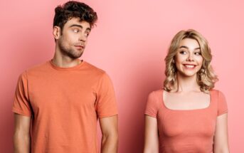 A man with curly dark hair in an orange t-shirt looks sideways at a smiling blonde woman in an orange top. Both are standing against a pink background, with the woman appearing cheerful and the man giving a skeptical glance.