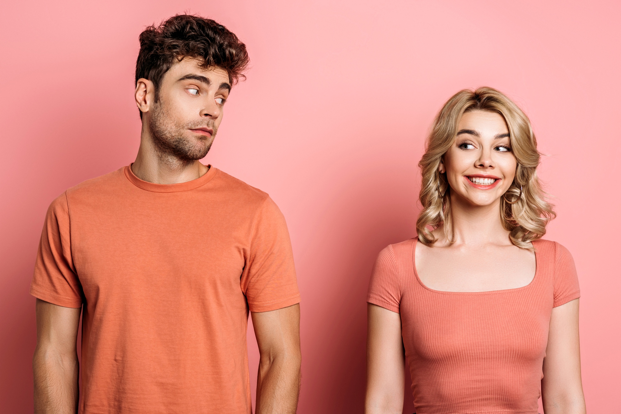 A man with curly dark hair in an orange t-shirt looks sideways at a smiling blonde woman in an orange top. Both are standing against a pink background, with the woman appearing cheerful and the man giving a skeptical glance.