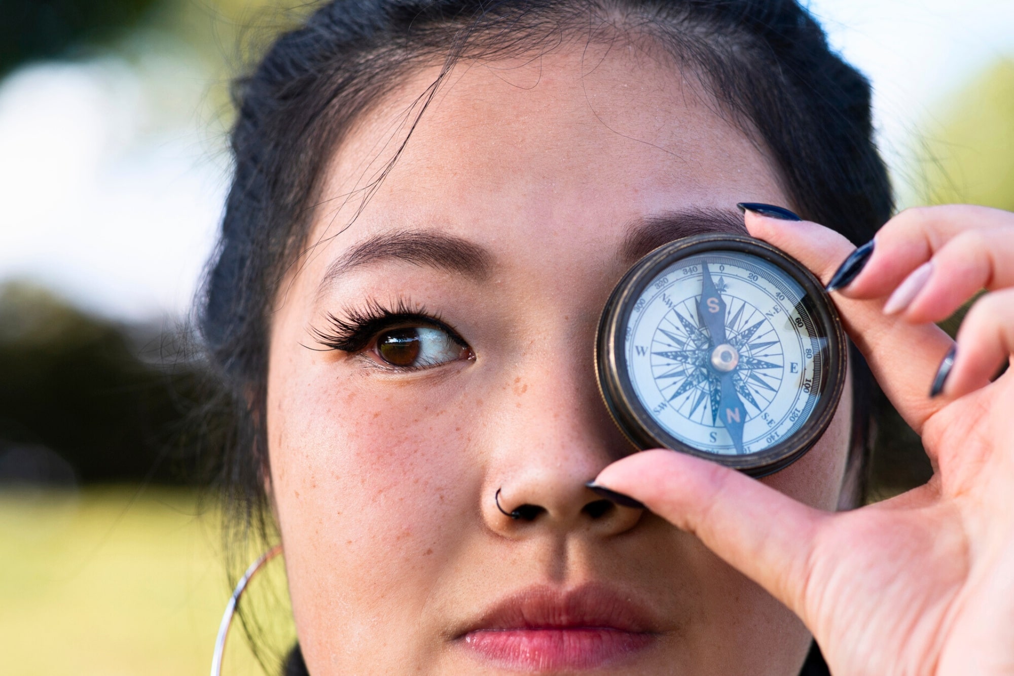 A person with dark hair holds a compass up to their right eye. They have a nose ring and large hoop earrings. The background is blurred greenery.