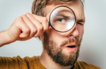 A man with brown hair and a beard holds a magnifying glass up to one eye, making it appear enlarged. He has a surprised expression and is wearing a mustard-colored shirt. The background is plain and light-colored.