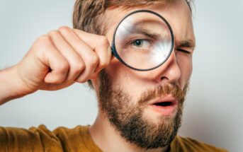 A man with brown hair and a beard holds a magnifying glass up to one eye, making it appear enlarged. He has a surprised expression and is wearing a mustard-colored shirt. The background is plain and light-colored.