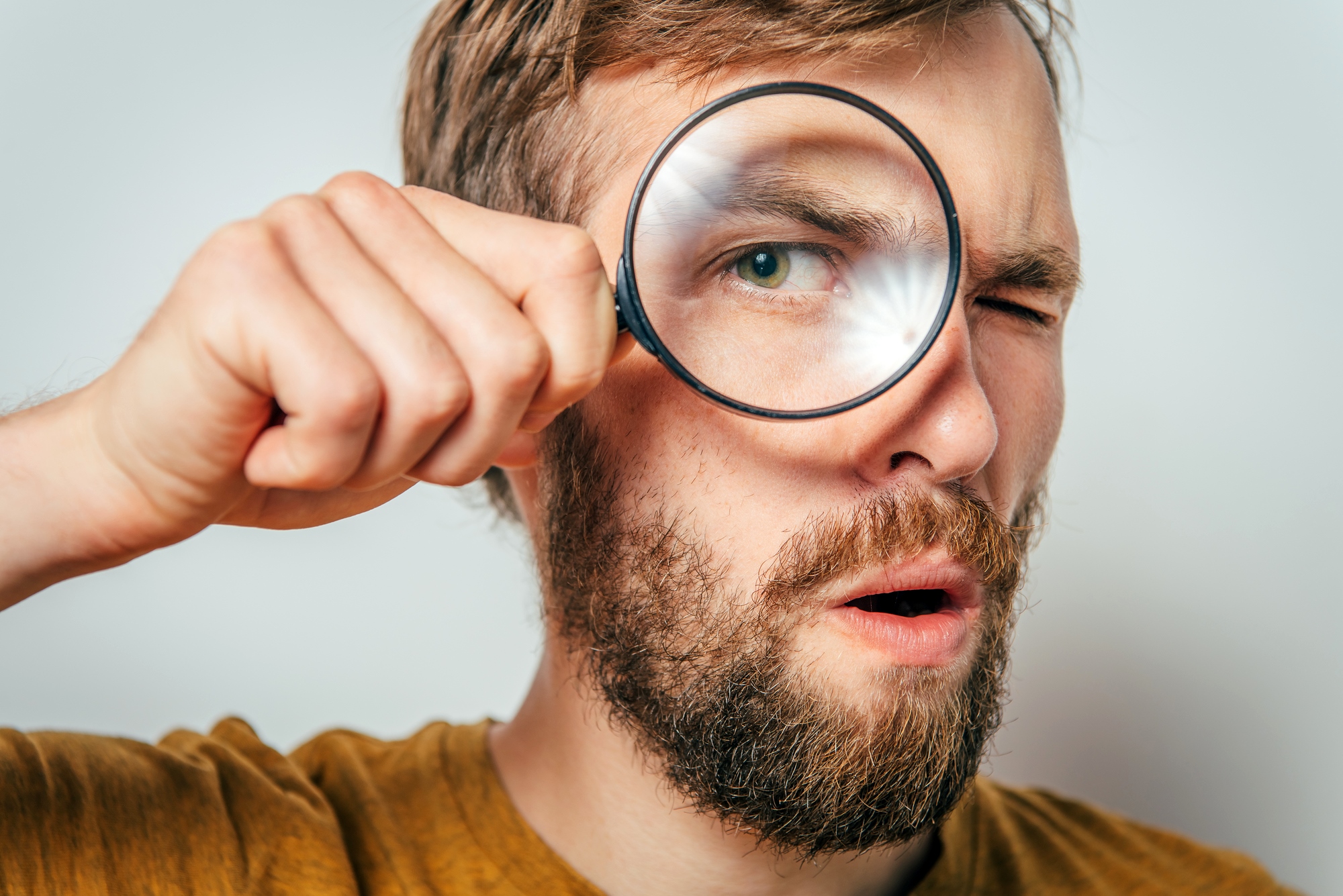 A man with brown hair and a beard holds a magnifying glass up to one eye, making it appear enlarged. He has a surprised expression and is wearing a mustard-colored shirt. The background is plain and light-colored.