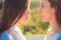 Two women with long brown hair face each other closely, standing in profile against a blurred backdrop of greenery. The sunlight highlights their faces and hair. Both are wearing white tops, creating a serene and contemplative mood.