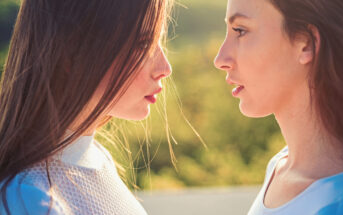 Two women with long brown hair face each other closely, standing in profile against a blurred backdrop of greenery. The sunlight highlights their faces and hair. Both are wearing white tops, creating a serene and contemplative mood.