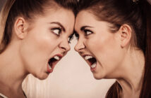 Two women with intense expressions are standing face-to-face, mouths open wide as if shouting at each other. Their brows are furrowed, and they appear to be in a heated argument. The background is neutral, keeping the focus on their emotional exchange.