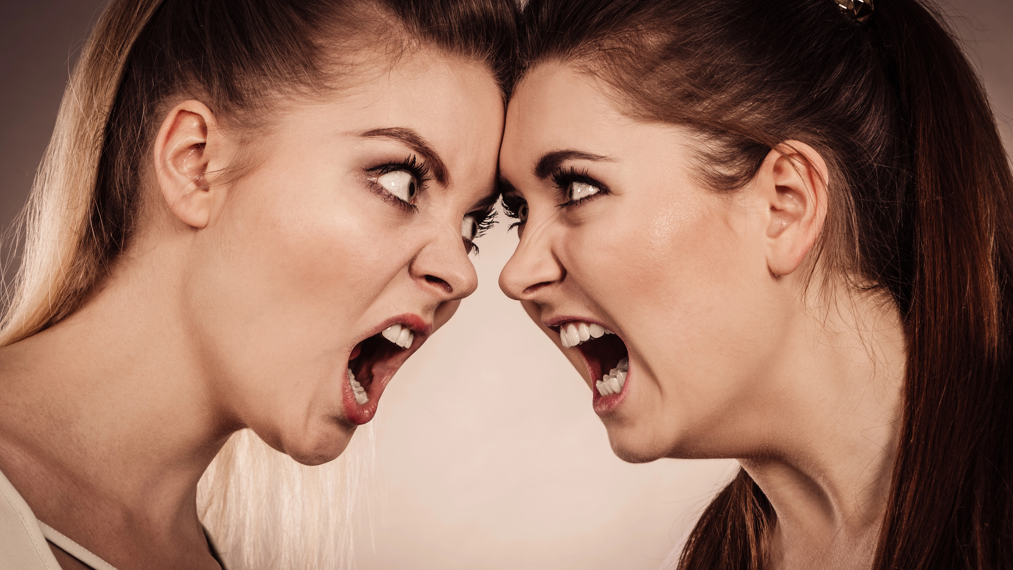 Two women with intense expressions are standing face-to-face, mouths open wide as if shouting at each other. Their brows are furrowed, and they appear to be in a heated argument. The background is neutral, keeping the focus on their emotional exchange.