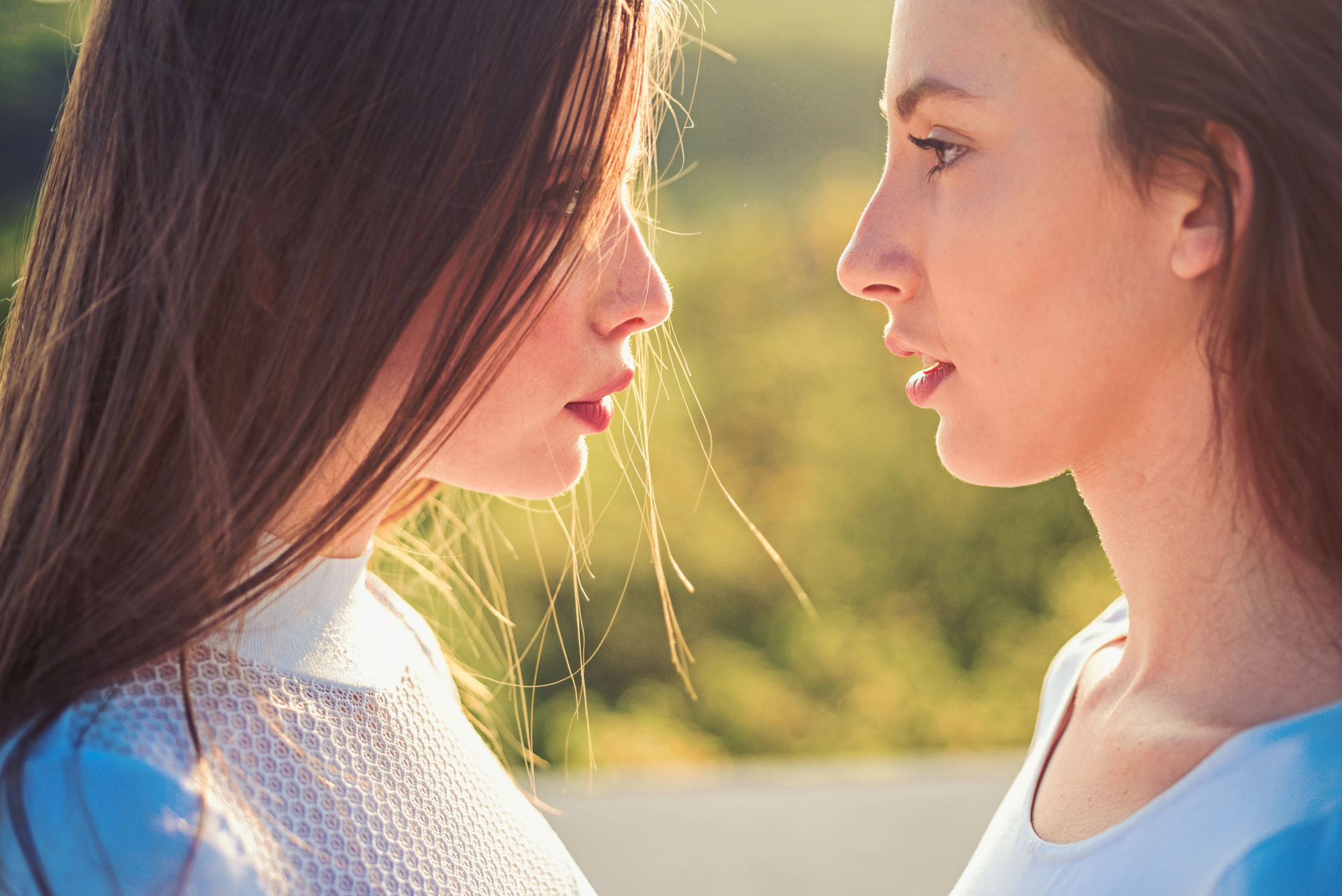 Two women with long brown hair face each other closely, standing in profile against a blurred backdrop of greenery. The sunlight highlights their faces and hair. Both are wearing white tops, creating a serene and contemplative mood.