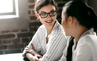 Two women are seated at a table, having a conversation. One woman with glasses and a striped shirt is smiling towards the other woman, who has her back to the camera and is wearing a white shirt. The setting appears to be a bright, sunny room with brick walls.