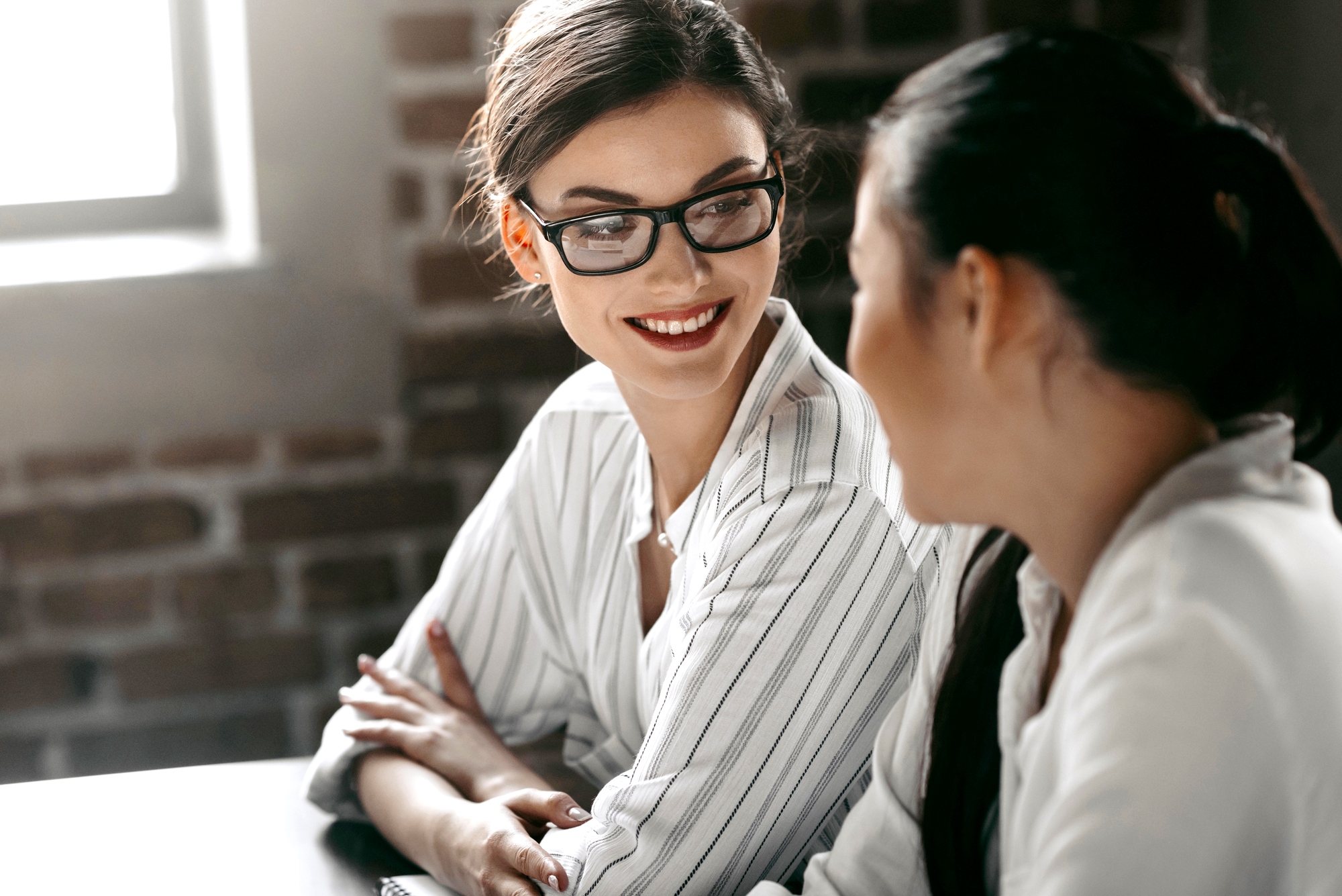 Two women are seated at a table, having a conversation. One woman with glasses and a striped shirt is smiling towards the other woman, who has her back to the camera and is wearing a white shirt. The setting appears to be a bright, sunny room with brick walls.