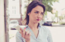 A woman stands outside holding a smartphone in one hand, appearing thoughtful. She is wearing a light blue button-up shirt and has shoulder-length dark hair. The background includes blurred buildings and greenery.