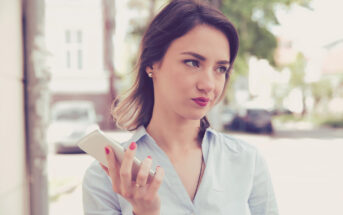 A woman stands outside holding a smartphone in one hand, appearing thoughtful. She is wearing a light blue button-up shirt and has shoulder-length dark hair. The background includes blurred buildings and greenery.