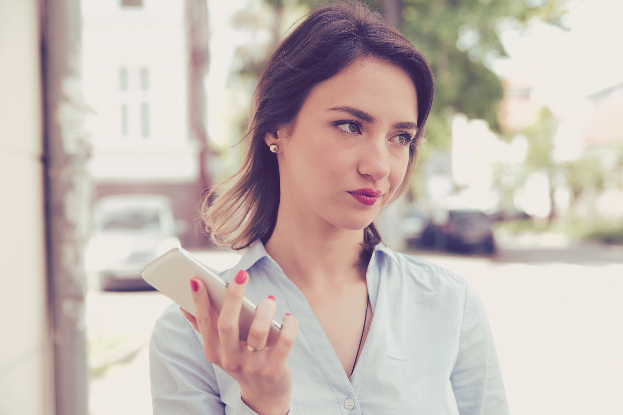 A woman stands outside holding a smartphone in one hand, appearing thoughtful. She is wearing a light blue button-up shirt and has shoulder-length dark hair. The background includes blurred buildings and greenery.