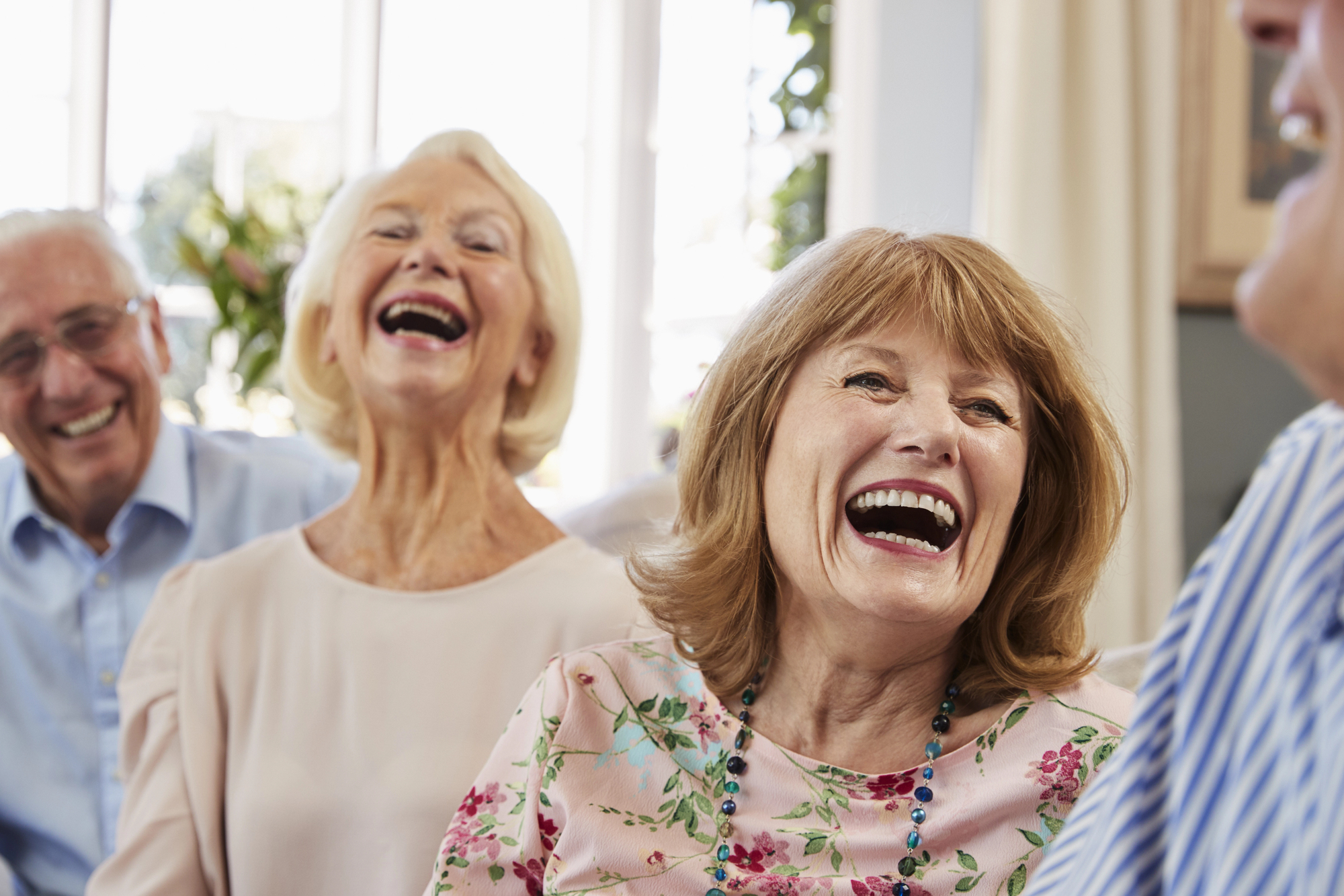 A group of elderly people sitting together in a bright room, laughing and enjoying each other's company. The focus is on two women in the foreground, both smiling widely, while two men in the background also share a laugh.