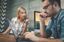 A woman and a man are working at a desk with laptops and desktop monitors in front of a textured gray wall. The woman is speaking seriously while the man, wearing glasses, looks thoughtful with his hand on his chin. A coffee cup and model airplanes are on the desk.