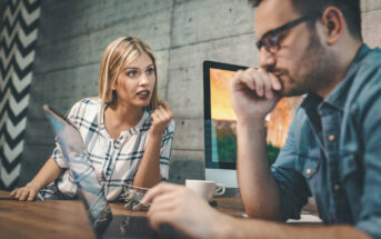 A woman and a man are working at a desk with laptops and desktop monitors in front of a textured gray wall. The woman is speaking seriously while the man, wearing glasses, looks thoughtful with his hand on his chin. A coffee cup and model airplanes are on the desk.