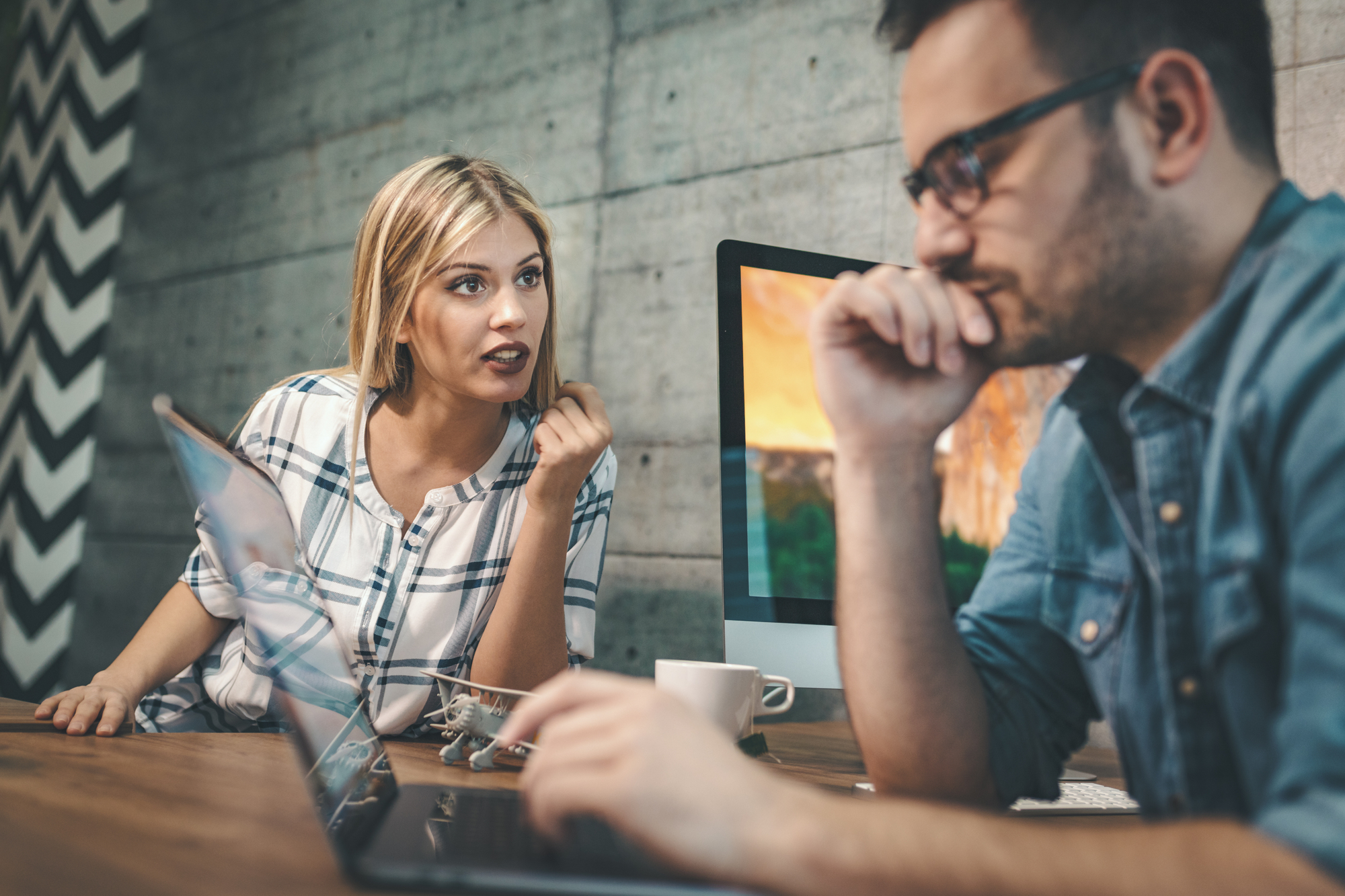 A woman and a man are working at a desk with laptops and desktop monitors in front of a textured gray wall. The woman is speaking seriously while the man, wearing glasses, looks thoughtful with his hand on his chin. A coffee cup and model airplanes are on the desk.