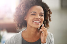 A smiling person with curly hair holds a phone to their ear. They are dressed in a grey top with a lighter grey cardigan. The background is blurred and brightly lit.