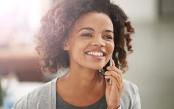 A smiling person with curly hair holds a phone to their ear. They are dressed in a grey top with a lighter grey cardigan. The background is blurred and brightly lit.