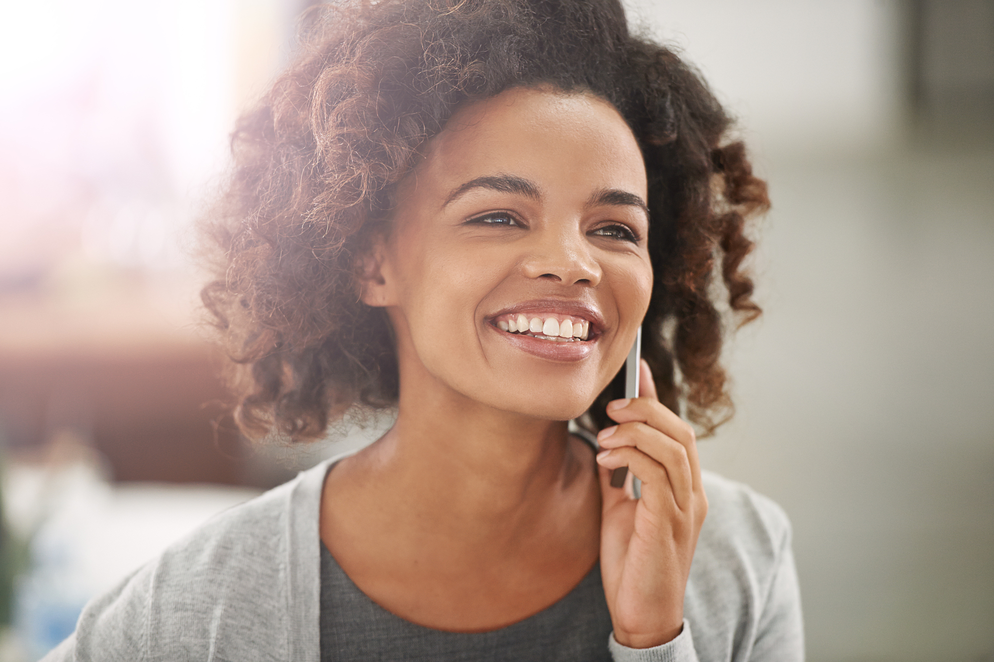 A smiling person with curly hair holds a phone to their ear. They are dressed in a grey top with a lighter grey cardigan. The background is blurred and brightly lit.