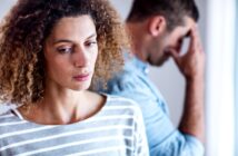 A woman with curly hair wearing a striped shirt looks down with a pensive expression. In the blurry background, a man in a blue shirt stands with his head down and hand covering his face, appearing distressed.