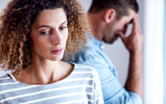 A woman with curly hair wearing a striped shirt looks down with a pensive expression. In the blurry background, a man in a blue shirt stands with his head down and hand covering his face, appearing distressed.