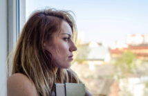 A woman with long, light brown hair stands by a window, gazing outside. She holds a closed book and has a contemplative expression on her face. The background features a blurry view of rooftops and trees.