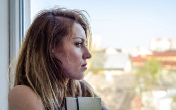 A woman with long, light brown hair stands by a window, gazing outside. She holds a closed book and has a contemplative expression on her face. The background features a blurry view of rooftops and trees.