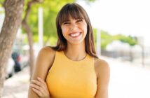 A woman with long brown hair and bangs smiles brightly while standing outdoors. She is wearing a sleeveless yellow top and has her arms crossed. The background includes blurred trees and a bright, sunny environment.