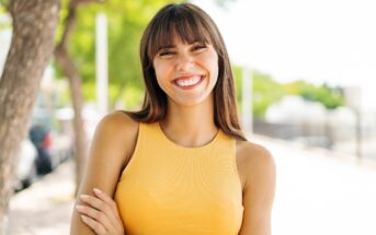 A woman with long brown hair and bangs smiles brightly while standing outdoors. She is wearing a sleeveless yellow top and has her arms crossed. The background includes blurred trees and a bright, sunny environment.