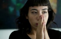 A person with short, dark hair and bangs is seen in a close-up, contemplative pose with hands pressed together as if in prayer or deep thought. The background is blurred, featuring dark and colorful tones. The individual appears to be indoors.