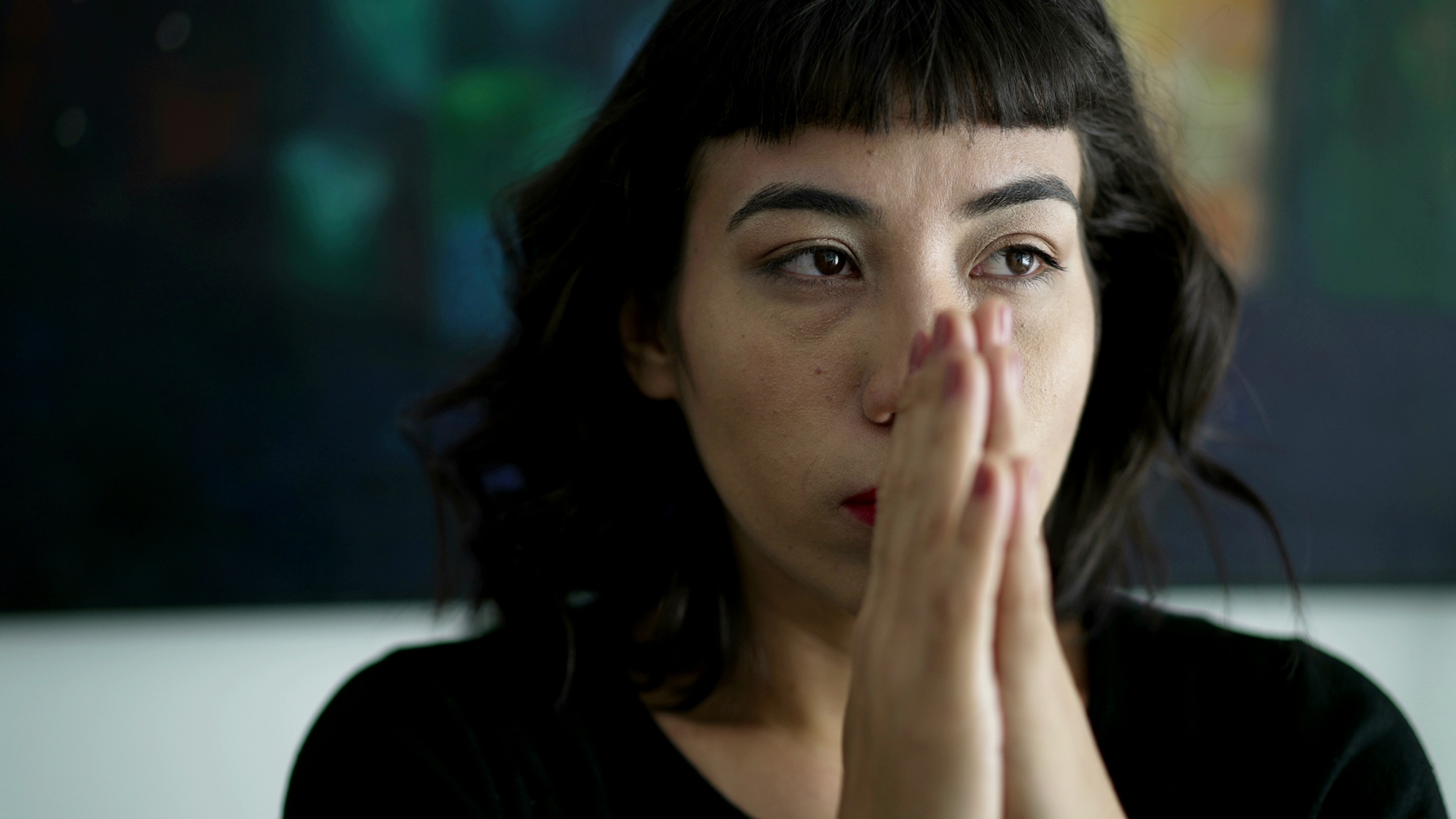 A person with short, dark hair and bangs is seen in a close-up, contemplative pose with hands pressed together as if in prayer or deep thought. The background is blurred, featuring dark and colorful tones. The individual appears to be indoors.