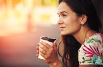 A woman with long brown hair, wearing a flowery shirt, is sitting outdoors in soft sunlight. She is holding a takeaway coffee cup with both hands and looking thoughtfully into the distance, with a slight smile on her face.