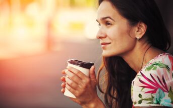 A woman with long brown hair, wearing a flowery shirt, is sitting outdoors in soft sunlight. She is holding a takeaway coffee cup with both hands and looking thoughtfully into the distance, with a slight smile on her face.