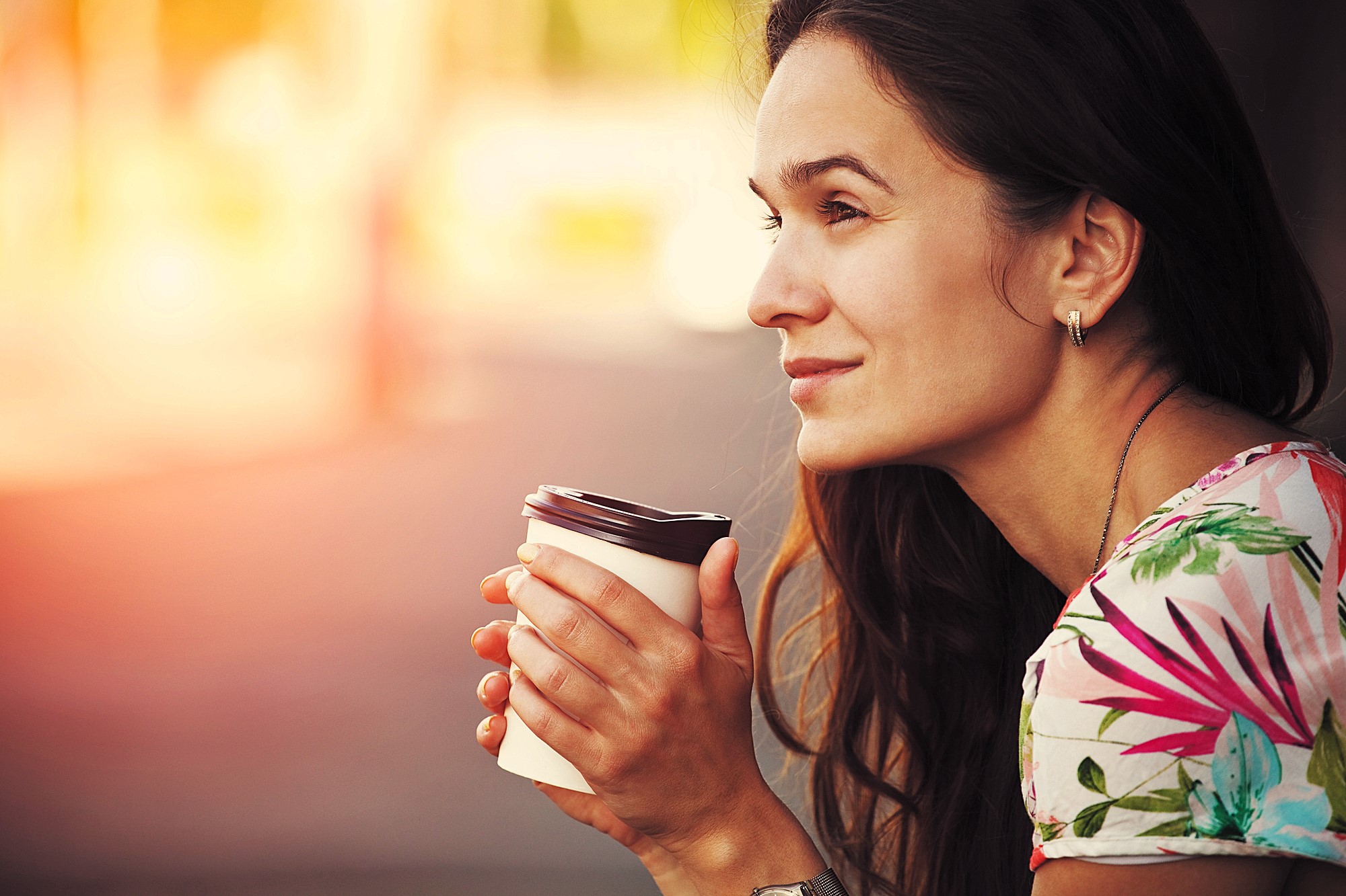 A woman with long brown hair, wearing a flowery shirt, is sitting outdoors in soft sunlight. She is holding a takeaway coffee cup with both hands and looking thoughtfully into the distance, with a slight smile on her face.