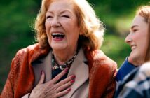 An elderly woman with blonde hair and a bright smile is laughing heartily with her hand on her chest. She is wearing a beige coat with an orange scarf. A younger woman next to her is smiling, looking at her. They are outdoors with a blurred green background.